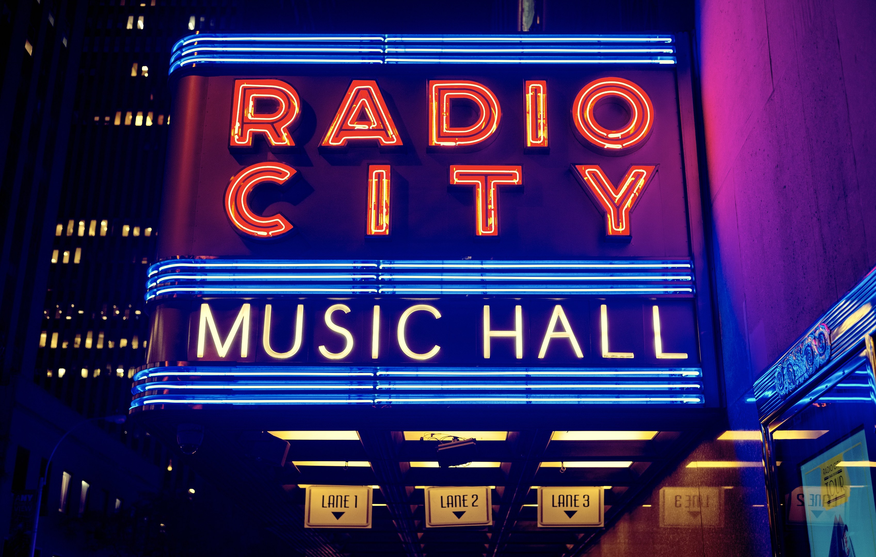 Radio City Music Hall in New York City, hell erleuchtet in der Nacht, umgeben von hohen Gebäuden, mit einem "Radio City"-Schild und mehreren kleineren Schildern auf der rechten Seite.