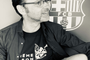 Ein Mann mit Brille und schwarzem Hemd sitzt vor einem schwarz-weißen FC Barcelona-Logo und lächelt.