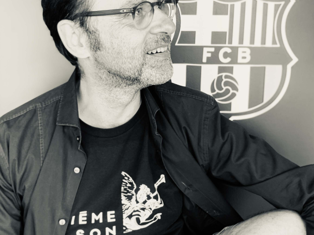 Ein Mann mit Brille und schwarzem Hemd sitzt vor einem schwarz-weißen FC Barcelona-Logo und lächelt.