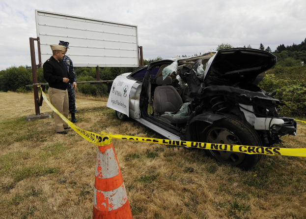 Ein Polizeibeamter in Uniform steht neben einem schwer beschädigten Auto am Straßenrand, mit eingedellter Motorhaube und zersplitterten Fenstern, während ein Verkehrskegel und ein Absperrband-marked Pole in der Nähe sind.