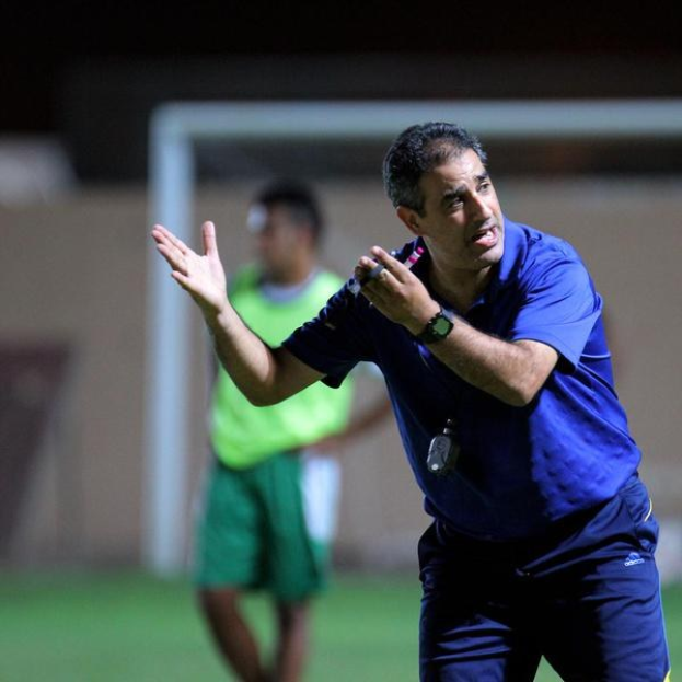 Ein Mann in einem blauen T-Shirt und blauen Hosen steht auf einem Fußballfeld und hält ein Mikrofon in der Hand, während er auf seine Spieler zeigt. Im Hintergrund ist eine Person in einem grünen Kleid, ein Torpfosten und eine Wand zu sehen.