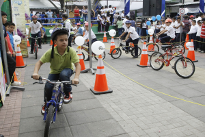 Kinder fahren Fahrräder auf einer Straße mit orangen Verkehrskegeln, einige tragen Helme, andere schauen zu; Bäume, Gebäude und ein klarer blauer Himmel im Hintergrund.