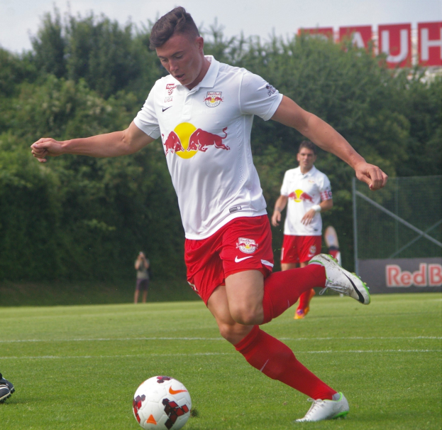 Ein professioneller Fußballspieler tritt einen Ball auf einem grünen Feld mit Bäumen und einem klaren blauen Himmel im Hintergrund neben einer Tafel mit der Aufschrift "RB Leipzig"