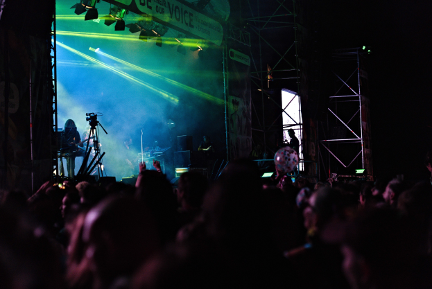 Eine Menge schaut Musikern auf einer hell erleuchteten Bühne mit Instrumenten, Mikrofonen und einem Banner bei einem Konzert zu.