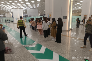 Menschen in einer Reihe an einem Flughafen-Sicherheits-Check-in, einige halten Taschen und Mobiltelefone, mit Säulen und Deckenlampen im Hintergrund.