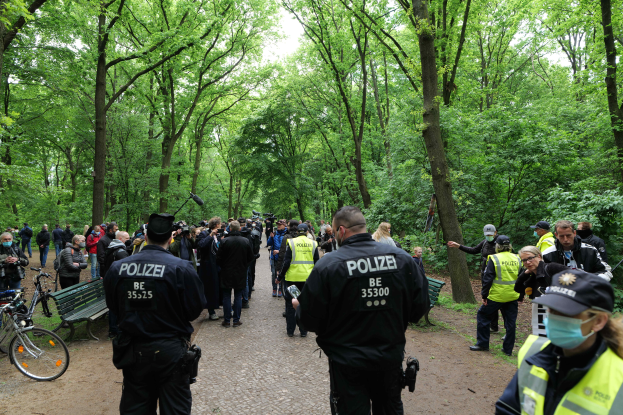 Eine Gruppe von Polizisten steht vor einer Menschenmenge, einige tragen Mützen und Masken, mit Fahrrädern und einer Bank im Vordergrund und Bäumen und Himmel im Hintergrund, wahrscheinlich während einer Anti-Terror-Demonstration in Berlin.