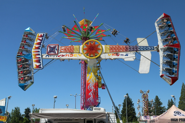 Ein Karussell auf einem Volksfest mit Menschen darauf, umgeben von Zelten, Bannern, Masten, Lichtern, Bäumen und anderen Gegenständen, mit dem Himmel im Hintergrund.