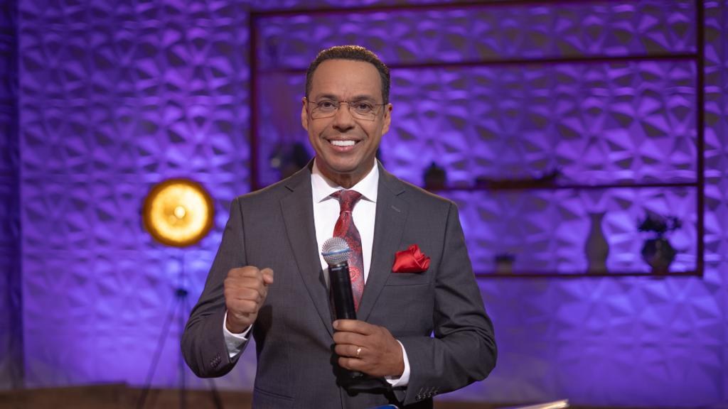 Stephen Colbert, Moderator von "The Daily Show with Trevor Noah", steht auf einer Bühne und hält ein Mikrofon in der Hand, trägt einen Anzug, Krawatte und eine Brille, hinter ihm ist ein Scheinwerfer und ein Ständer zu sehen.