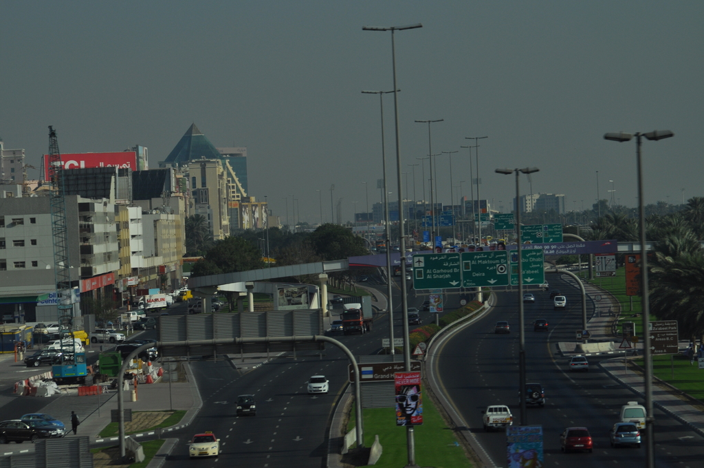 Eine Luftaufnahme einer Stadt mit Gebäuden, Türmen, Kraftfahrzeugen, Werbetafeln, Schildern, Straßen, Bäumen, Straßenlaternen, Menschen, Verkehrskegeln und Informationsschildern unter einem klaren Himmel.