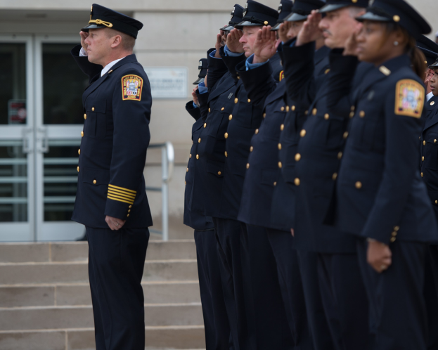Eine Gruppe von Polizisten in Uniform und mit Mützen, die in Formation vor einem Gebäude mit Glastüren und Treppen salutieren.