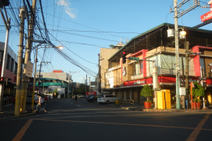 Eine Stadtstraßenszene mit Autos, Fußgängern, Strommasten, Ampeln, Gebäuden mit Schildern, Blumentöpfen und einem bewölkten Himmel.