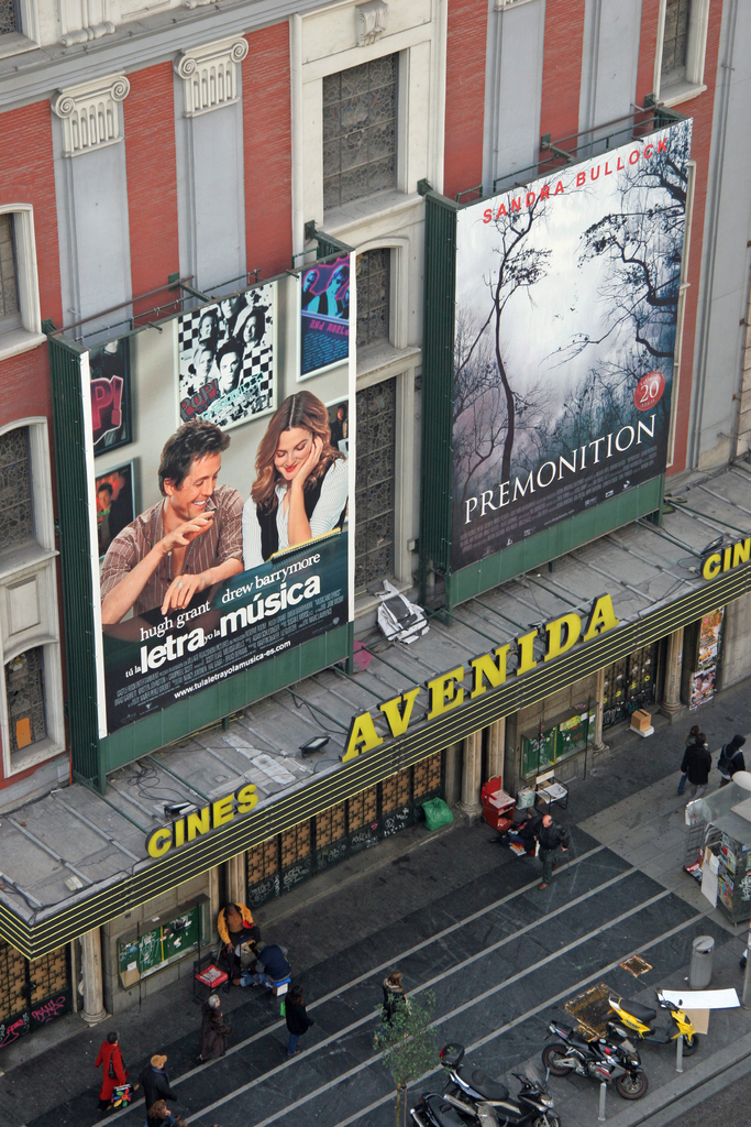 Ein Gebäude mit zwei Werbetafeln in der Mitte, Menschen, die gehen und auf dem Boden sitzen, und mehrere Motorräder, die in der Nähe einer Wand auf der rechten Seite geparkt sind.