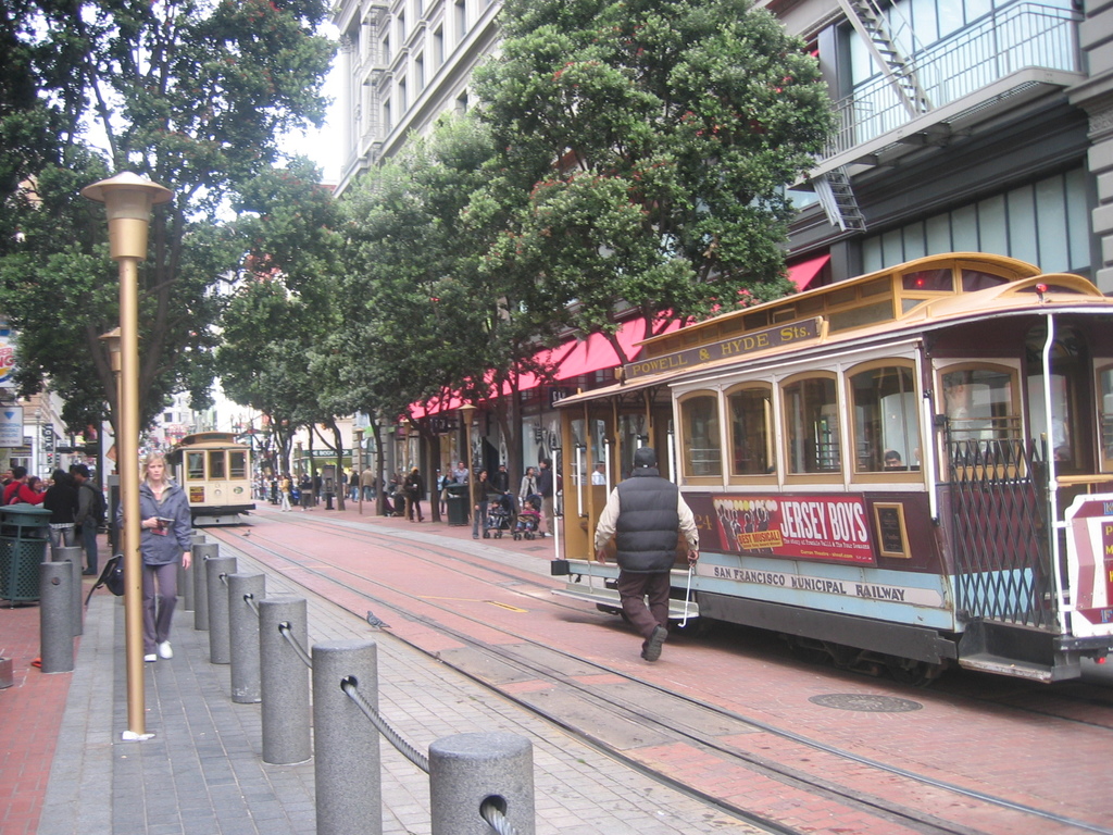 Eisenbahnschienen verlaufen entlang einer Straße mit darauf fahrenden Zügen, einer davon mit Text, und einer Person in der Nähe; kleine Pfosten mit Seilen und Straßenlaternen säumen die linke Seite, während Gebäude mit Leitern auf der rechten Seite stehen und Bäume und viele Menschen im Hintergrund zu sehen sind.