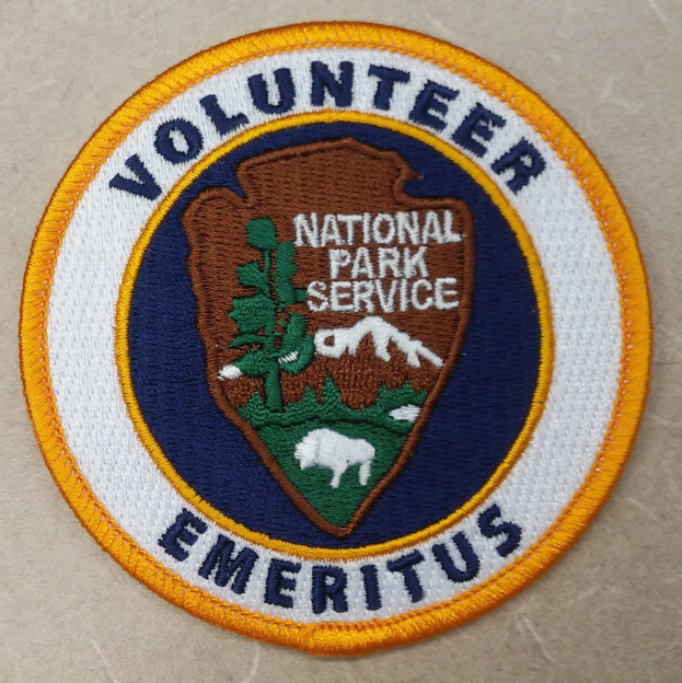Runder Patch mit "Volunteer National Park Service Emeritus" in fetter schwarzer Schrift auf weißem Hintergrund, umrandet von einem blauen Rand und einem gelben Kreis.