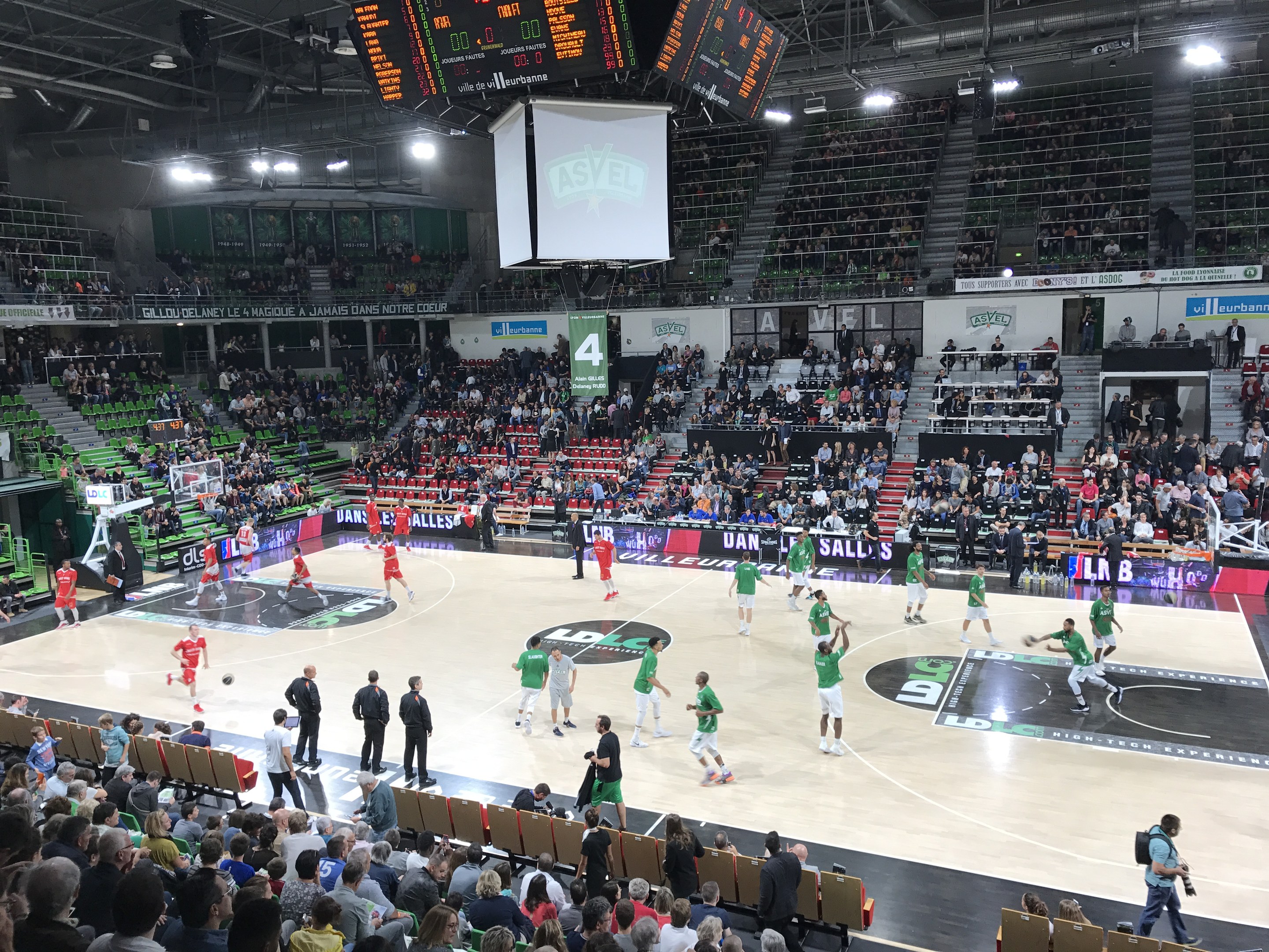Basketballspiel in einer vollen Halle mit Zuschauern, Anzeigetafeln und Stadionbeleuchtung.