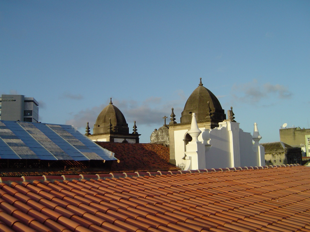 Stadtansicht mit mehreren Gebäuden im Vordergrund und einem klaren blauen Himmel im Hintergrund, mit Solarpanelen auf einem Dach, die den Einsatz erneuerbarer Energien anzeigen.