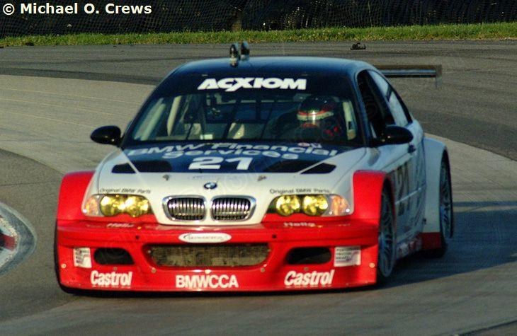 Eine Person in einem Helm fährt einen BMW M3 GT3 Rennauto auf einer von Gras und einem Maschendrahtzaun umgebenen Rennstrecke.
