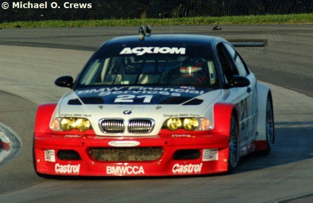 Eine Person in einem Helm fährt einen BMW M3 GT3 Rennauto auf einer von Gras und einem Maschendrahtzaun umgebenen Rennstrecke.