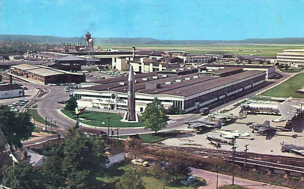 Altes Schwarz-Weiß-Foto eines Flughafens mit geparkten Flugzeugen, Gebäuden, Bäumen, Straßen, Fahrzeugen, Strommasten, Gras, Bahnschienen, Türmen, fernen Hügeln und einem klaren Himmel, mit Text am unteren Rand.