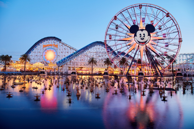 Eine lebendige Dämmerungsszene im Disneyland California Adventure mit einem riesigen Mickey-Maus-Rad im Vordergrund, einer Achterbahn im Hintergrund und Booten auf dem Wasser, mit einem farbenfrohen Himmel und silhouettenartigen Bäumen.
