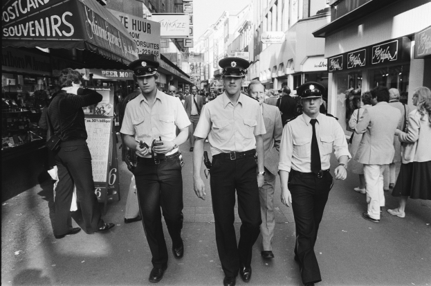 Drei Polizisten in Uniform gehen eine belebte Stadtstraße mit hohen Gebäuden, Geschäften und Passanten entlang.