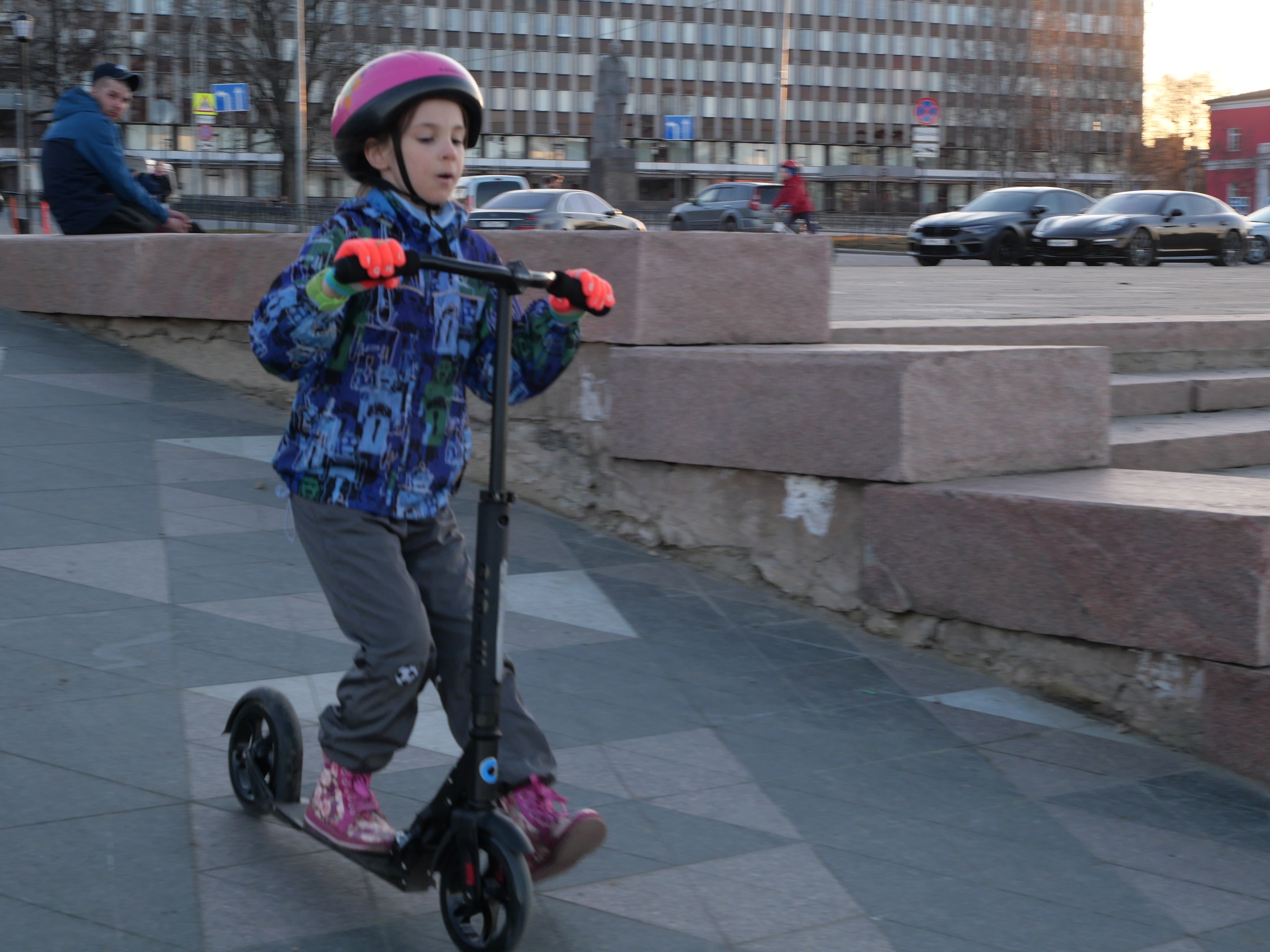 Ein junger Junge mit Helm und Handschuhen fährt auf einem Gehweg mit Treppen, Fahrzeugen, Menschen, Bäumen, Pfählen, Brettern, Gebäuden und einem klaren blauen Himmel im Hintergrund.