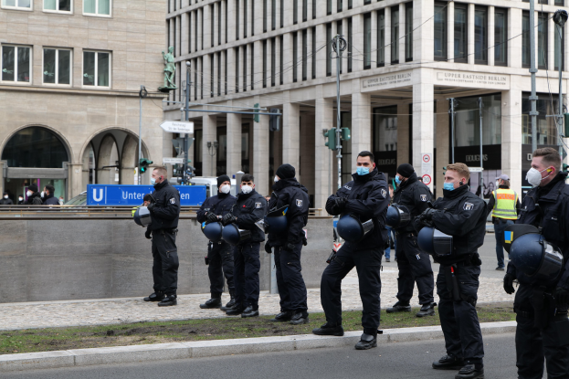 Polizisten in schwarzen Uniformen und Masken vor einem Berliner Gebäude mit Glasfenstern, Säulen und grasbewachsenem Boden.
