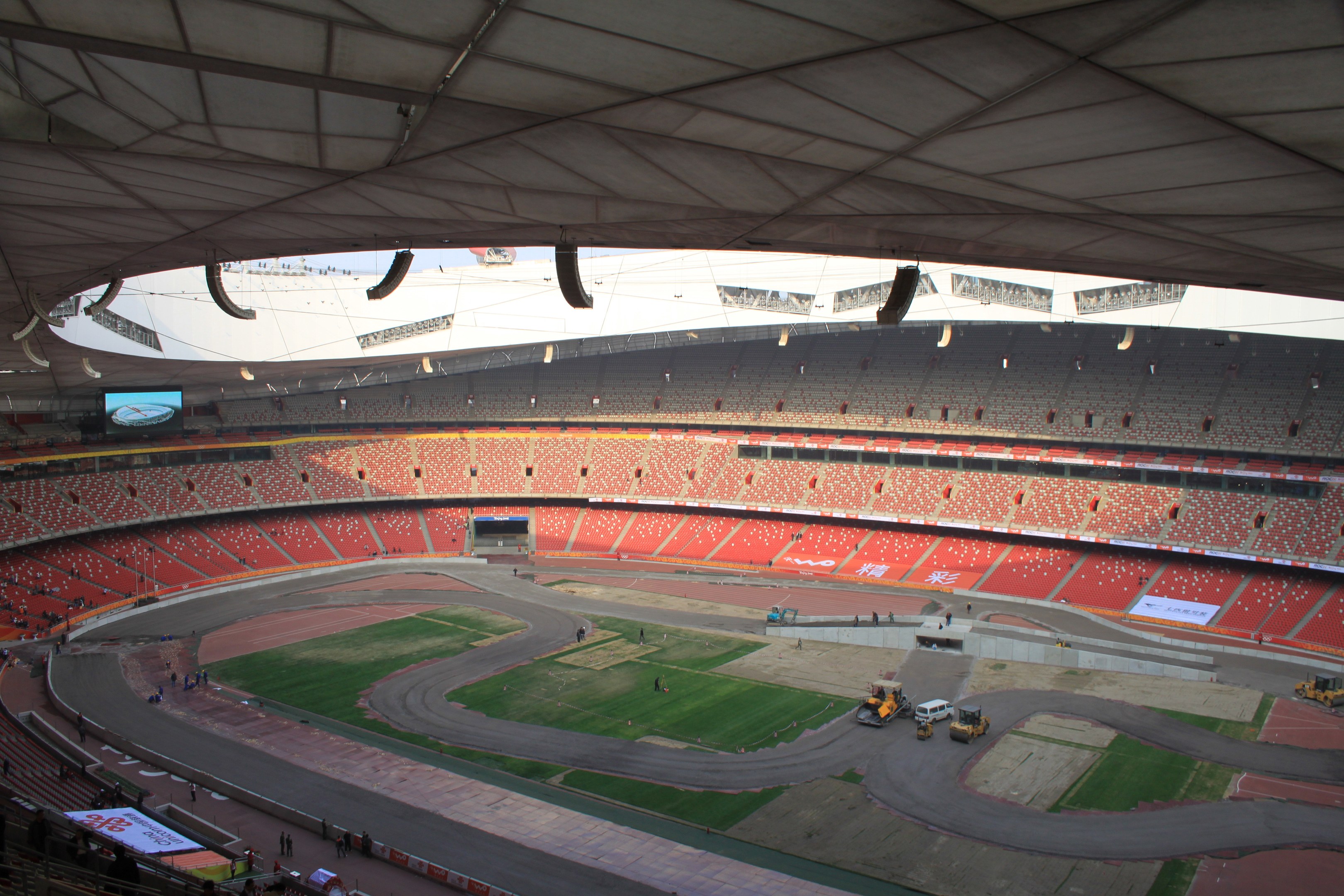 Großes Stadion voller Zuschauer, umgeben von Fahrzeugen, Gras, Texttafeln, Geländern, einem Bildschirm und Deckenbeleuchtung.