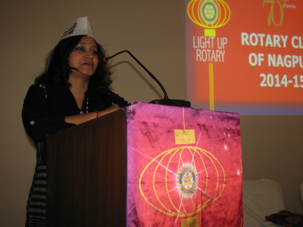 Eine Frau in einer Mütze und einem Kleid steht an einem Podium mit einem Mikrofon und einem daran befestigten Tuch und spricht vor einer Wandanzeige.