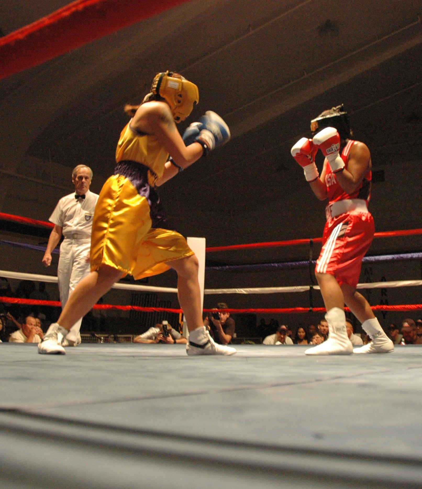 Zwei Boxer kämpfen in einem Ring, während ein Referee beobachtet, umgeben von Fotografen und Zuschauern unter einer sichtbaren Decke.