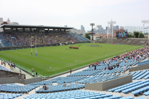 Ein großes Stadion voller Zuschauer bei einem Fussballspiel, mit sitzenden und stehenden Zuschauern, Pfosten, Lichtern, einem Bildschirm, Bäumen, Gebäuden und einem klaren blauen Himmel.
