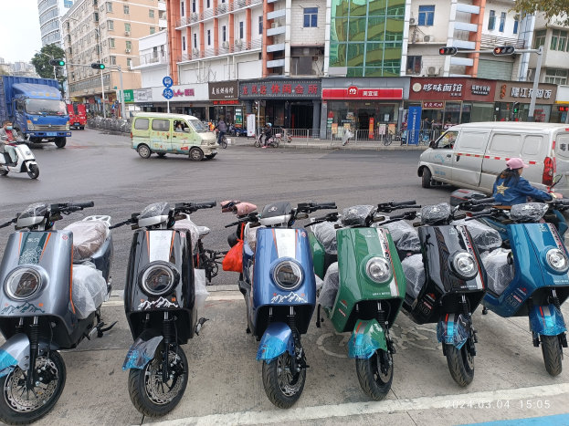 Eine Reihe von E-Scootern auf einer Straße geparkt, mit einer Person in einer Mütze davor, vor einem Hintergrund aus Gebäuden, Verkehrsampeln, Bäumen und einem klaren Himmel.