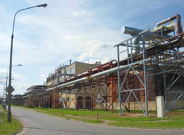 Große Fabrik mit umfangreichen Rohrleitungen, umgeben von Gebäuden, Straßeninfrastruktur, Grünflächen und einem bewölkten Himmel.