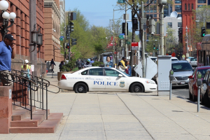 Eine belebte Stadtstraße mit einem geparkten Polizeiwagen, fahrenden Fahrzeugen einschließlich eines Busses und eines Autos, Fußgängern auf dem Gehweg, Gebäuden mit Fenstern, Laternen, Geländern, Bäumen und einem klaren blauen Himmel.