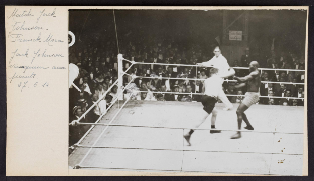 Schwarzes und weißes Foto von zwei Boxern, Jack Johnson und Frank Morgan, in einem Boxring mit Zuschauern und einer Wand im Hintergrund.