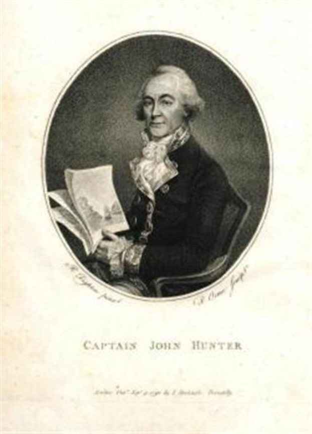 Captain John Hunter, der eine blaue Anzug, ein weißes Hemd und eine rote Krawatte trägt, sitzt in einem Stuhl und liest ein Buch, mit einem ernsten Gesichtsausdruck. Der Text "Captain John Hunter" wird unten im Bild angezeigt.