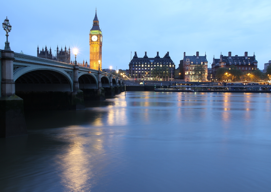 Ein See mit einer Brücke auf der linken Seite, ein Uhrenturm hinter der Brücke, Gebäude mit Lichtern im Hintergrund und Bäume vor den Gebäuden unter einem Himmel.