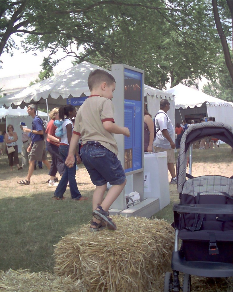 Eine Szene mit mehreren Zelten und Menschen, die darum herumlaufen, ein Anzeigetafel davor, ein Kind, das auf trockenem Gras daneben spielt, und Bäume auf beiden Seiten.