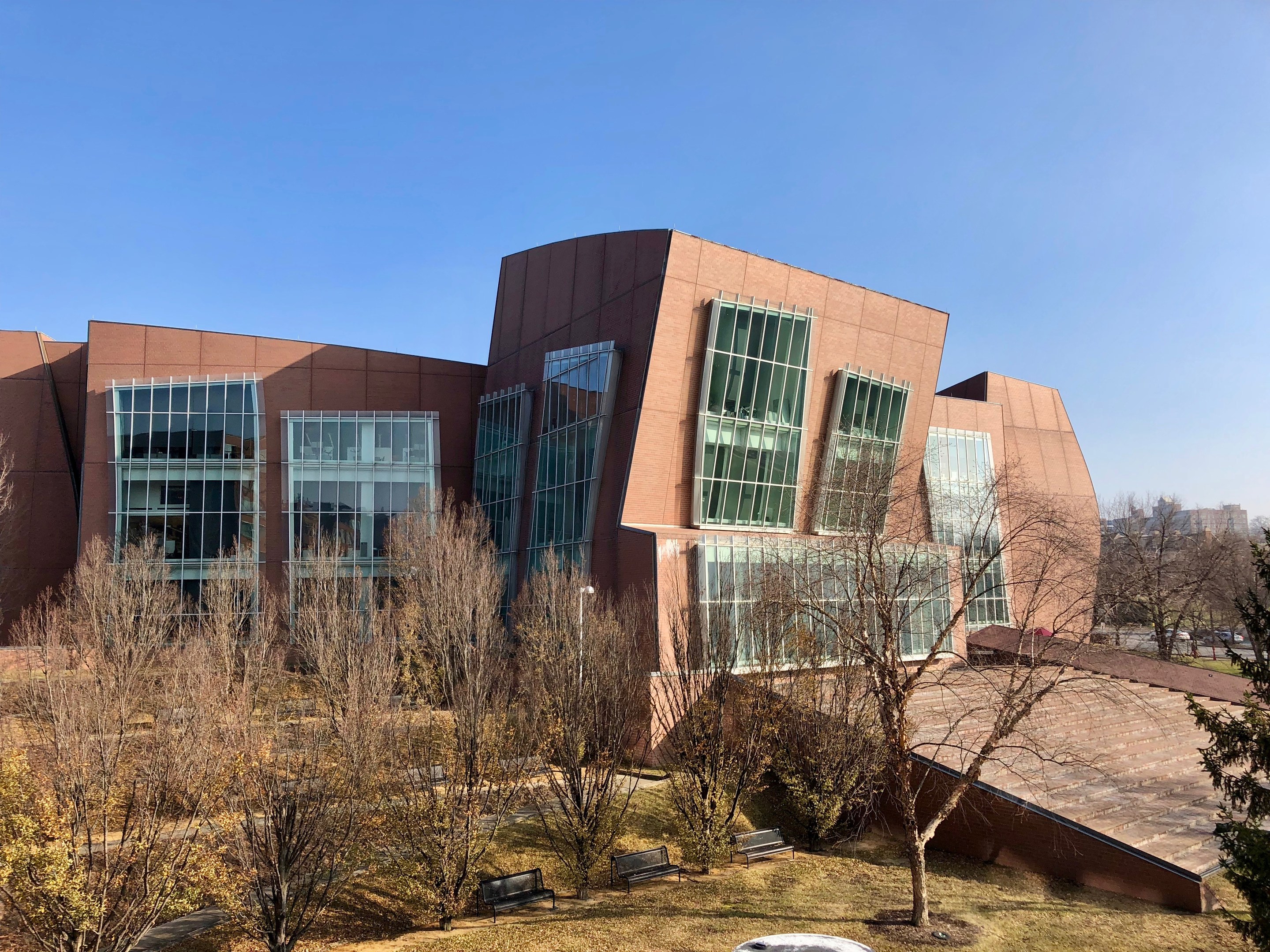 Großes Gebäude mit zahlreichen Fenstern, umgeben von Bäumen, Bänken, Gras und einem Lichtmast, vor anderen Gebäuden und einem klaren blauen Himmel, als der neue Campus der University of Illinois at Chicago identifiziert.