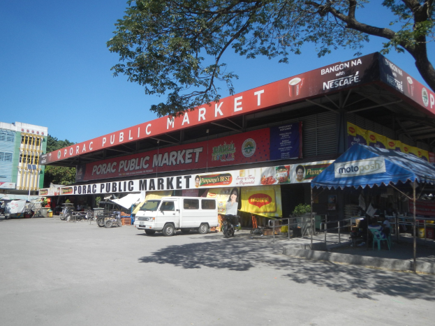 Ein ├Âffentlicher Markt mit einem vor ihm geparkten Van, umgeben von Geb├Ąuden, Gesch├Ąften, Namenstafeln, Kraftfahrzeugen auf der Stra├če, Personen unter einem Zelt sitzend, Gel├Ąndern, B├Ąumen und einem klaren blauen Himmel.