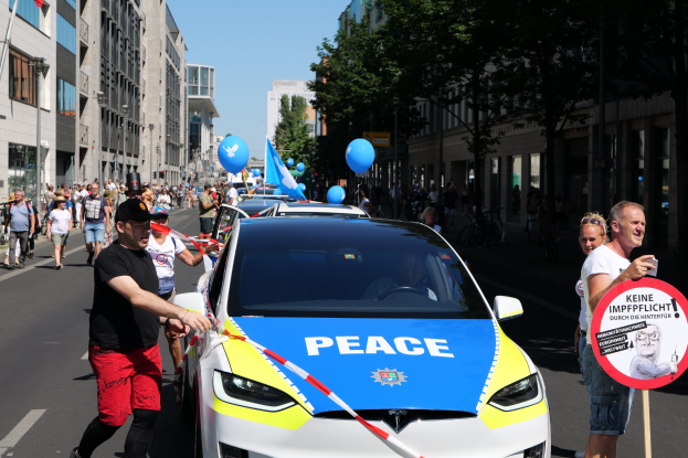 Eine Gruppe von Menschen geht neben einem Polizeiwagen her, der mit blauen, gelben und roten Schleifen geschmückt ist und auf dem das Wort 'Frieden' steht, vor einer Kulisse aus Gebäuden, Bäumen, Fahnen und einem klaren blauen Himmel.
