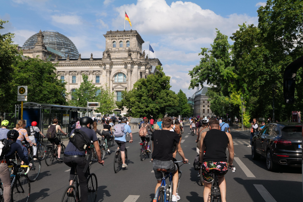 Gruppe von Menschen, die auf Fahrrädern eine straße mit Bäumen in Berlin, Deutschland, entlangfahren, mit Gebäuden und einer Bushaltestelle, unter einem bewölktem Himmel und einer Flagge auf einem Gebäude.