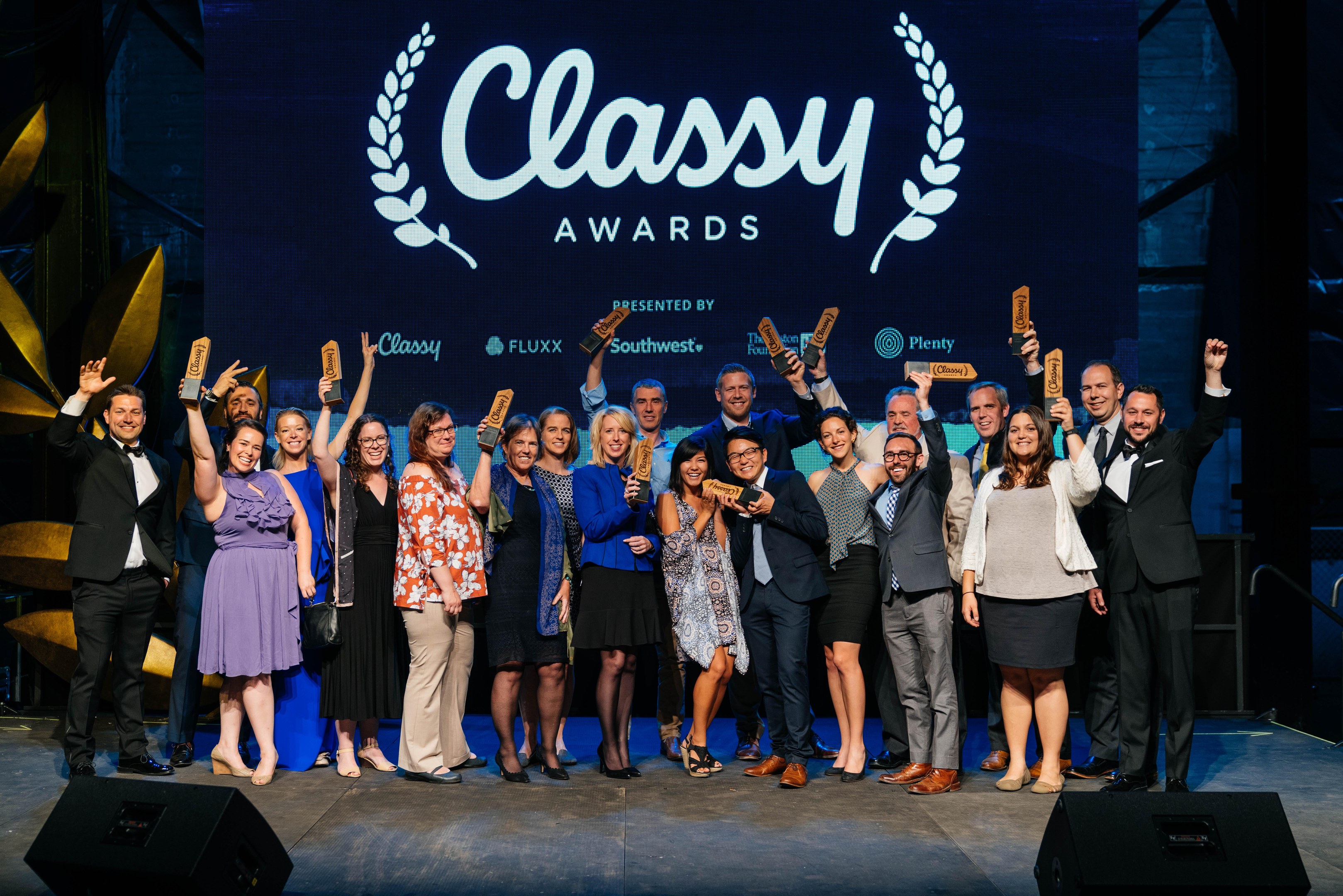 Gruppe von Menschen auf einer Bühne mit Auszeichnungen, mit Lautsprechern darunter und einem Bildschirm im Hintergrund, der "Classy Awards 2019" anzeigt.
