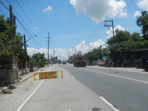 Eine Straße mit Fahrzeugen, ein "Verlangsamen Sie die Schule"-Schild, Strommasten mit Drähten, Bäume, Straßenlaternen und ein bewölkter Himmel.