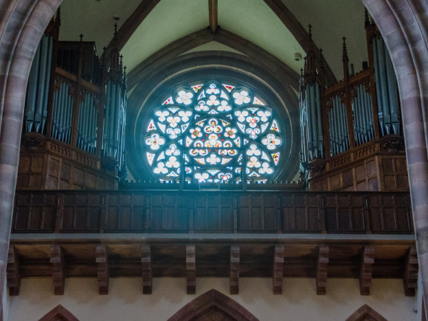 Innenansicht einer Kirche mit einem zentralen Buntglasfenster, das von Holzgeländern und Säulen umgeben ist, mit verstreuten Gegenständen und einer Wand unten.