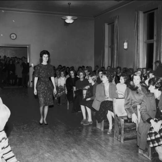 Eine Frau in einem schwarzen und weißen Kleid läuft über einen Laufsteg, während eine Zuschauermenge eine Modenschau zum Feiern der Frauenwahlrecht beobachten.