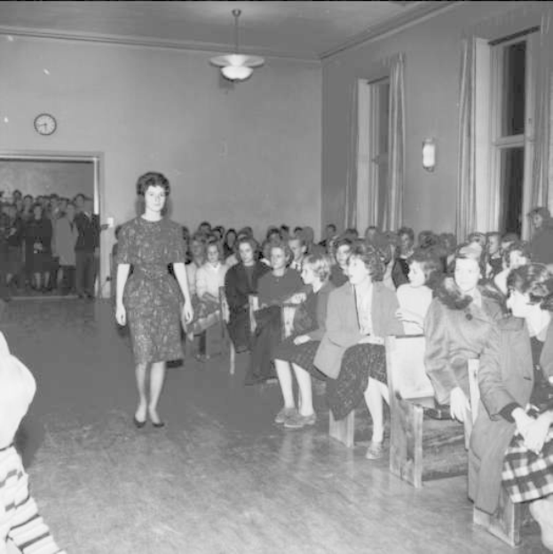 Eine Frau in einem schwarzen und weißen Kleid läuft über einen Laufsteg, während eine Zuschauermenge eine Modenschau zum Feiern der Frauenwahlrecht beobachten.