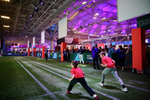 Zwei junge Jungs spielen Fußball auf einem Feld, umgeben von einer Gruppe von Menschen, die auf dem Boden stehen, Flaschen und Gegenstände rechts, ein Zaun, Banner mit Text, ein Anzeigebildschirm, Metallrahmen, ein Dach mit Deckenleuchten und Metallpfosten im Hintergrund.