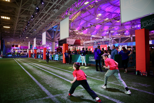 Zwei junge Jungs spielen Fußball auf einem Feld, umgeben von einer Gruppe von Menschen, die auf dem Boden stehen, Flaschen und Gegenstände rechts, ein Zaun, Banner mit Text, ein Anzeigebildschirm, Metallrahmen, ein Dach mit Deckenleuchten und Metallpfosten im Hintergrund.
