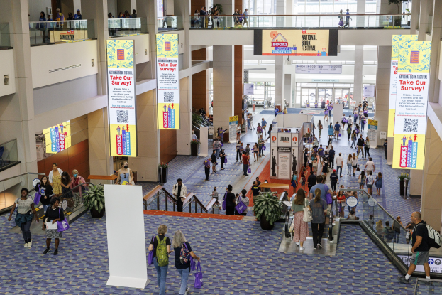 Eine große Menschenmenge, die durch ein großes Gebäude mit Bannern, Topfpflanzen, Rolltreppen, Geländern, Säulen, bedruckten Plakaten, Glastüren und anderen Gegenständen geht, wobei einige Taschen tragen.